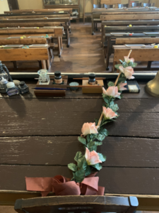 Mit Blumen verzierter Rohrstock auf einem Lehrerpult. Im Hintergrund sind die Holzbänke der Schüler zu sehen. Üblicherweise tuen dies die Schüler am Geburtstag des Lehrers.